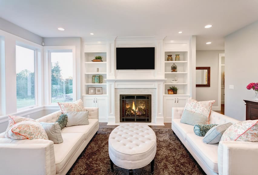 White ottoman, TV above fireplace and brown carpet