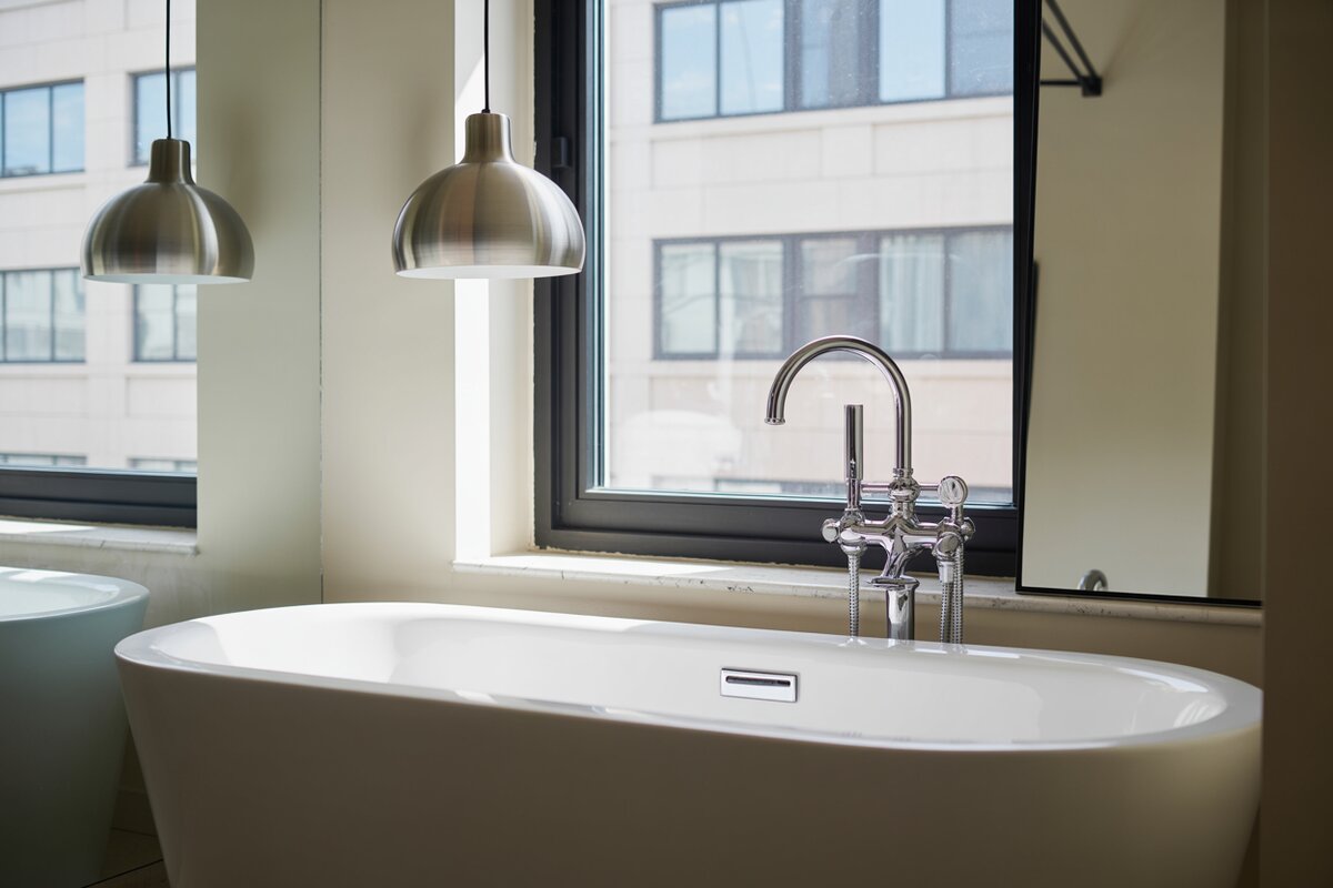 Two brushed metal dome pendants beside a window over a white freestanding tub