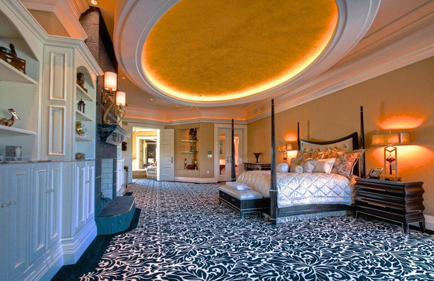 Large bedroom in upscale home with circular ceiling feature, white built-ins, fireplace and patterned carpet