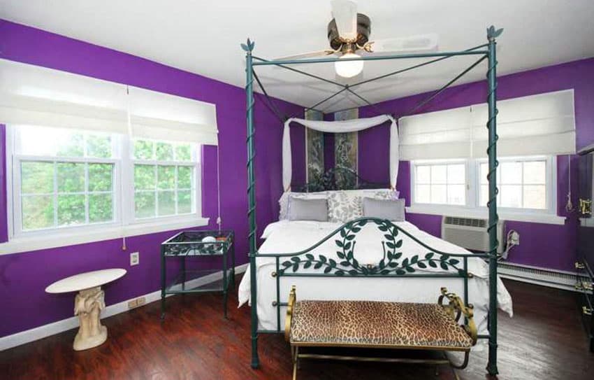 Bright purple bedroom with four poster bed and dark wood floors and white picture windows