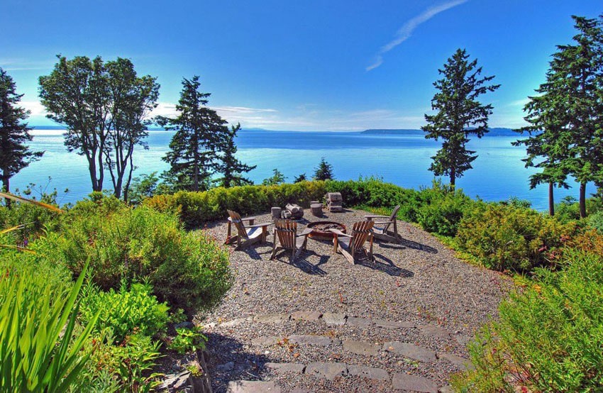 A breathtaking view of a lake with Adirondack chairs enciricling a pit