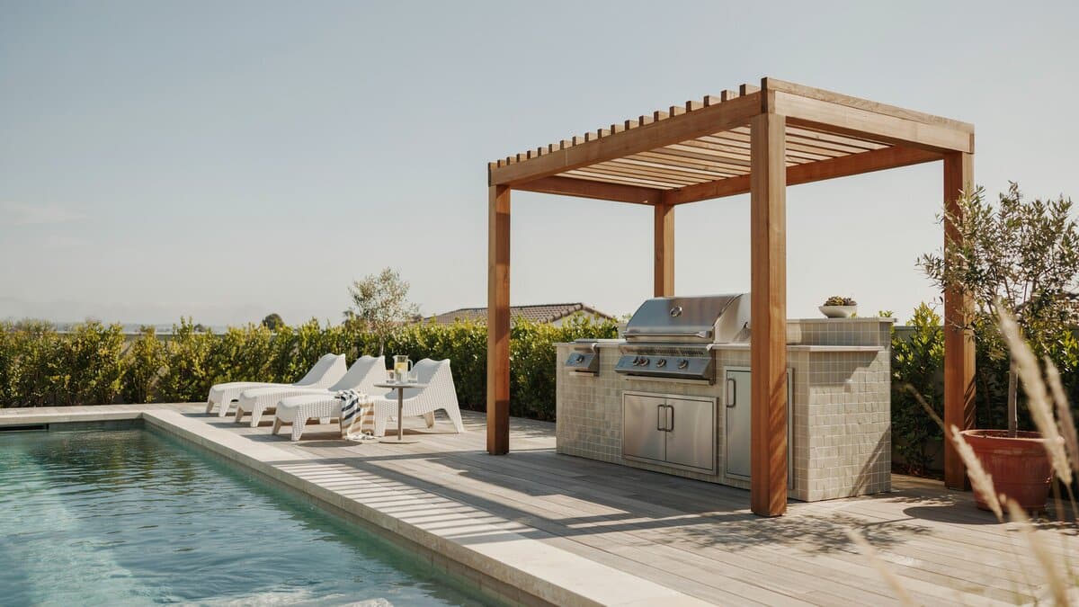 Standalone stainless barbecue island under a gazebo beside a swimming pool