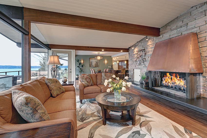 Rustic waterfront living room with large copper fireplace, rock accent wall and wood beams