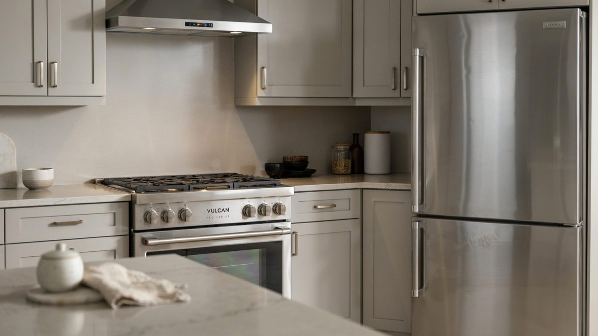 Stainless gas range, vent hood and refrigerator set into pale grey shaker cabinetry