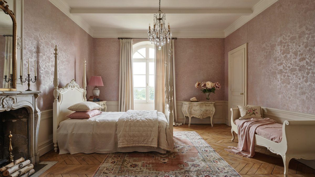 Pink walled bedroom with carved white bed, stone fireplace and crystal chandelier