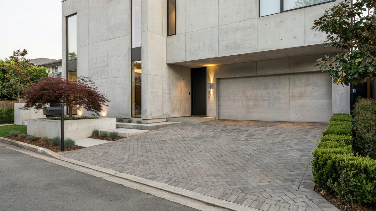 Herringbone paver drive tucked beside a board formed concrete house with a red maple