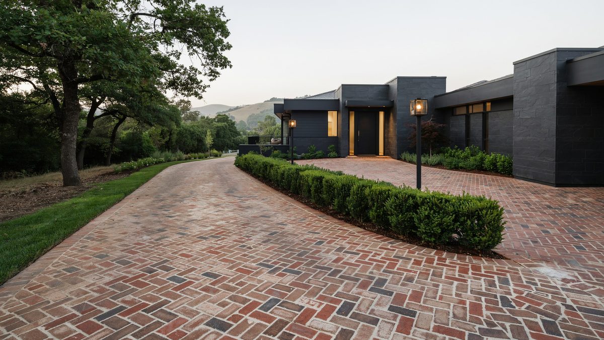 Red and charcoal brick drive winding past clipped hedges to a low dark modern house