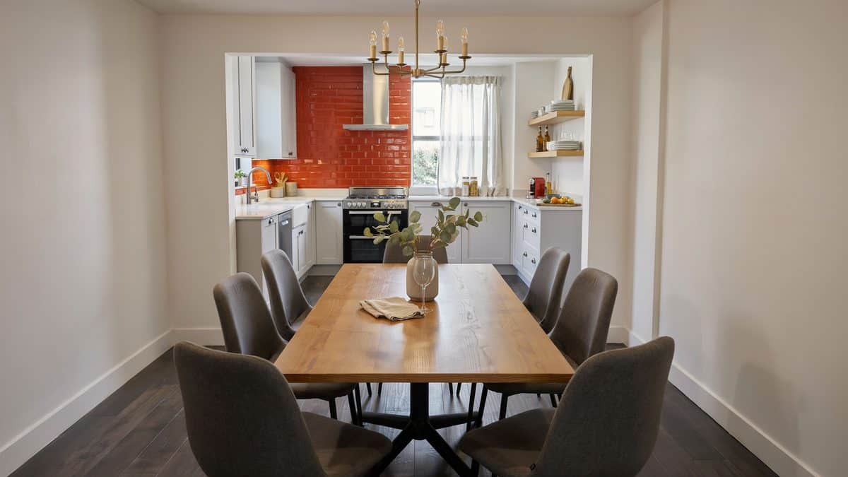 Oak table and gray chairs in a narrow room facing an orange backsplash kitchen