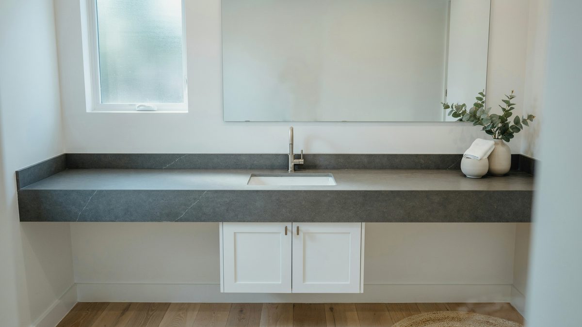 Long dark gray slate vanity counter above white cabinets under a small window