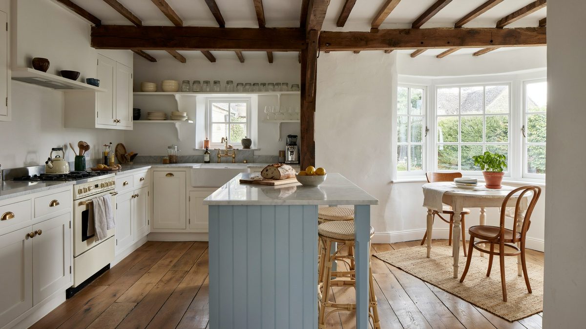 A soft blue island sits under rough timber beams beside a bay window table