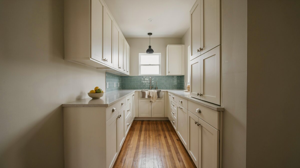 Narrow galley with aquamarine glass tile and warm teak flooring leading to a window