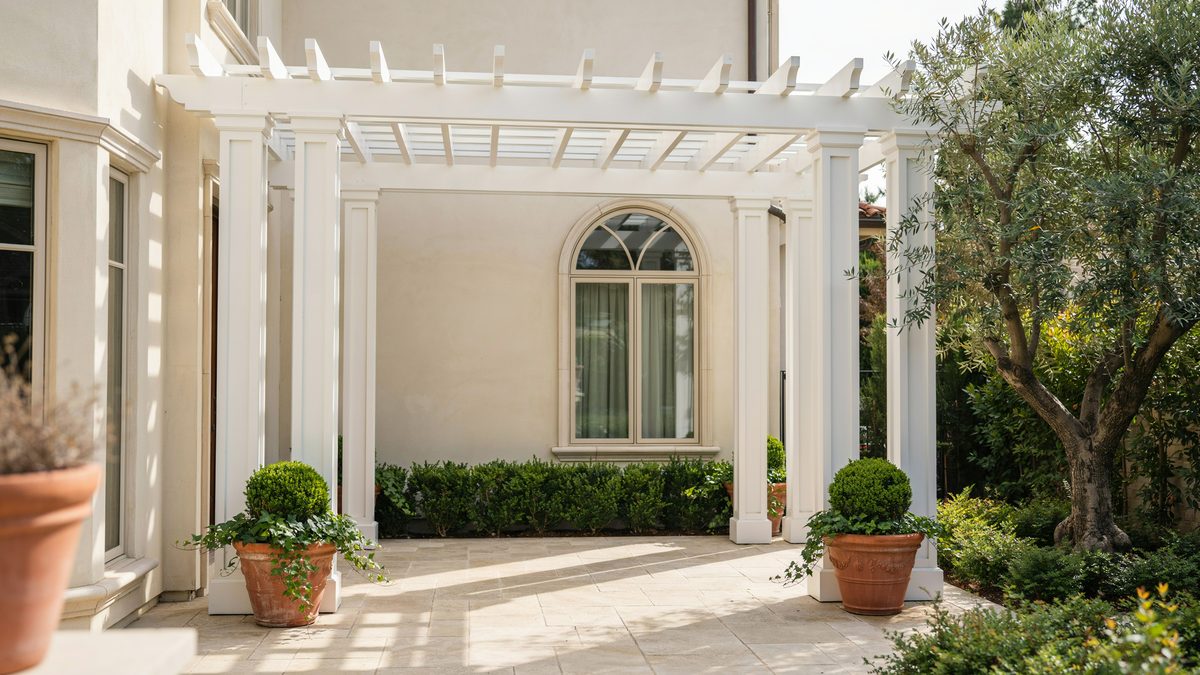 White neoclassical pergola attached to a stucco wall with an arched window
