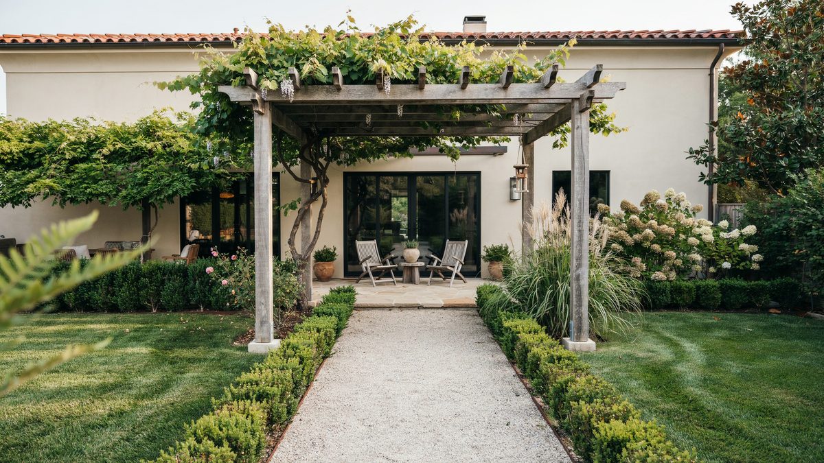 Grapevine covered pergola framing a gravel path toward a stucco house