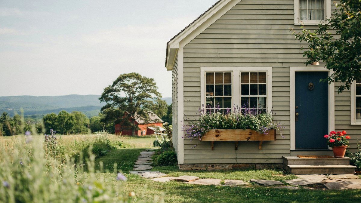 Wooden window box of purple flowers on a grey shingled farmhouse with rocking chair