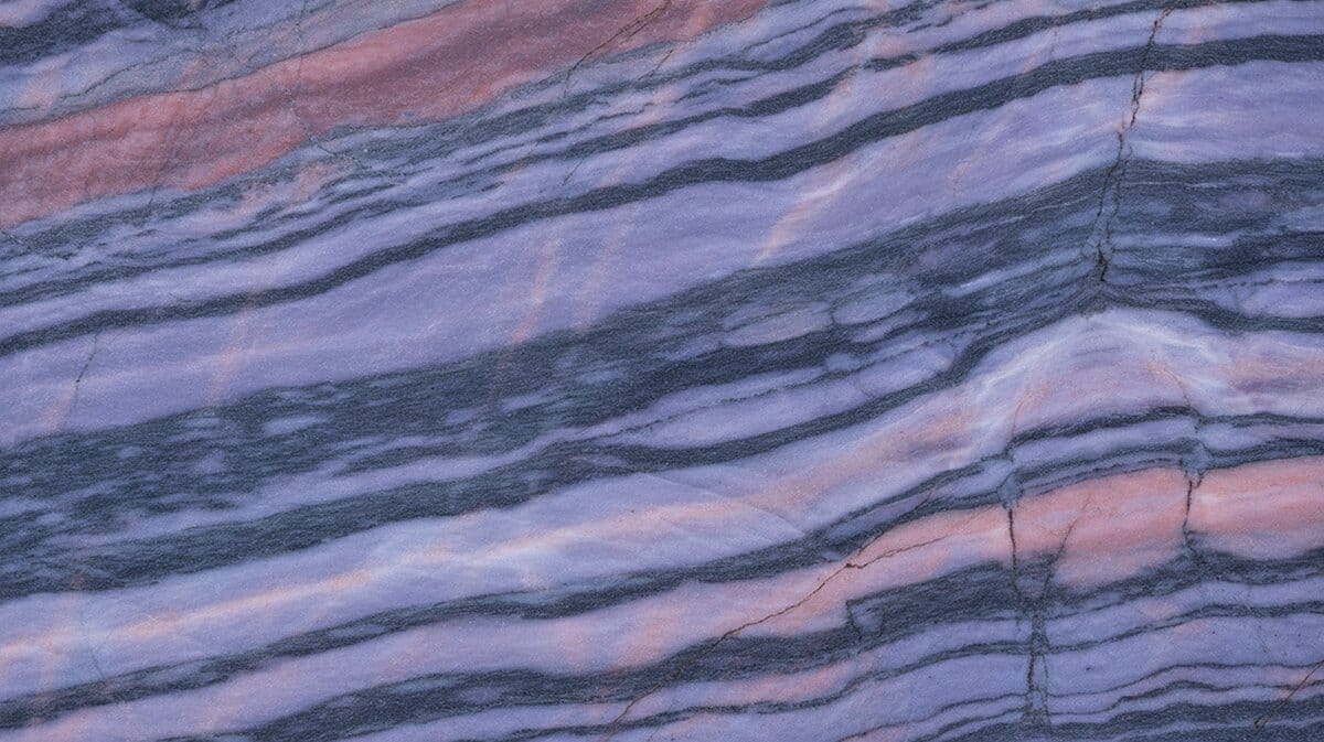 Aquarella Tycoon Blue granite slab with layered waves of purple lavender and rose pink sweeping across dark gray banded stone