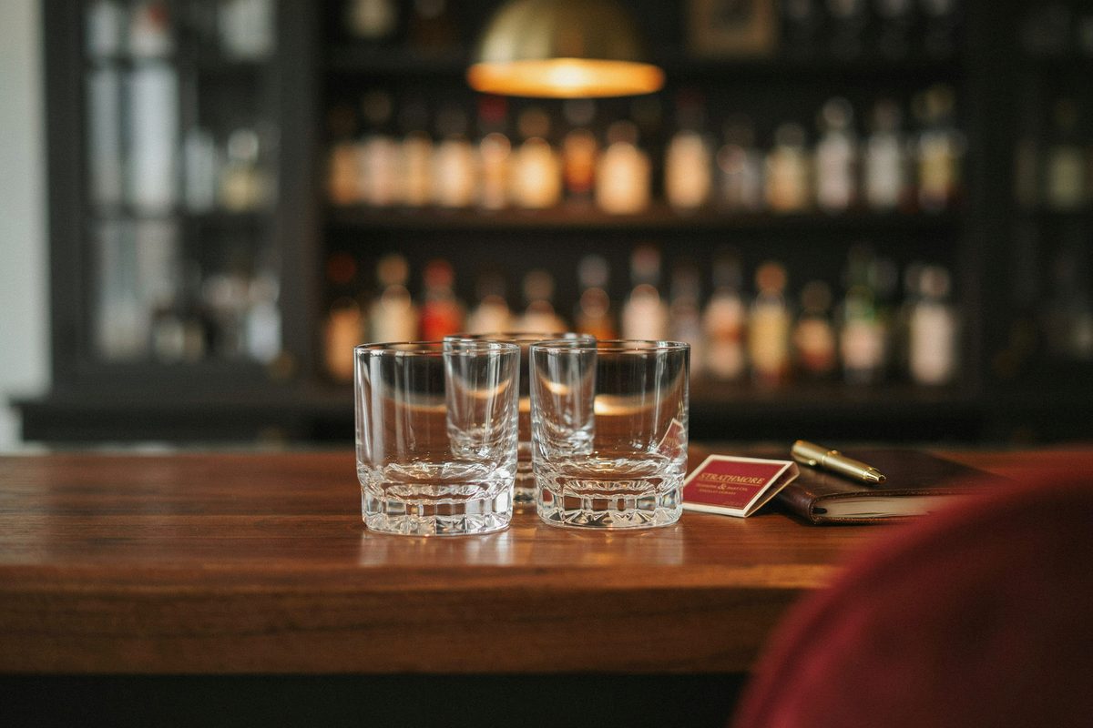 Three cut crystal whisky tumblers on a walnut counter before backlit bottles