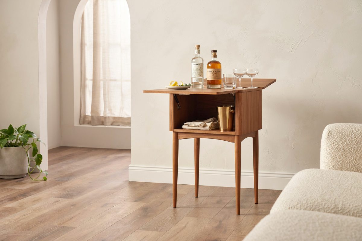 Walnut drop leaf portable bar cabinet holding bottles beside a sheer curtain