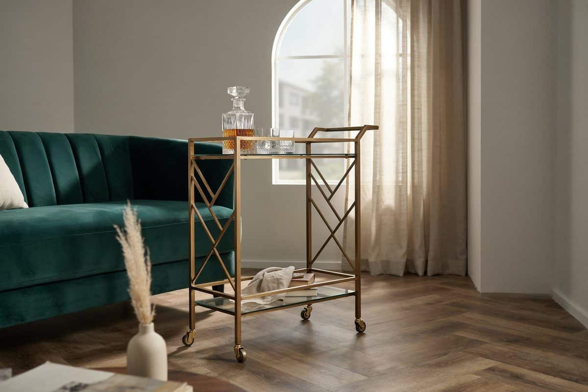 Brass and glass bar cart parked beside a teal velvet sofa