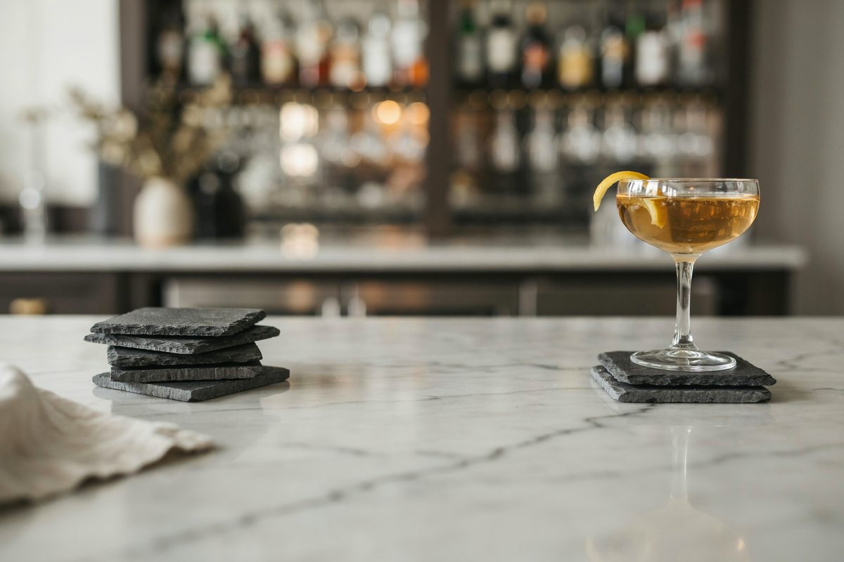 Square slate coasters stacked on marble next to a garnished coupe cocktail