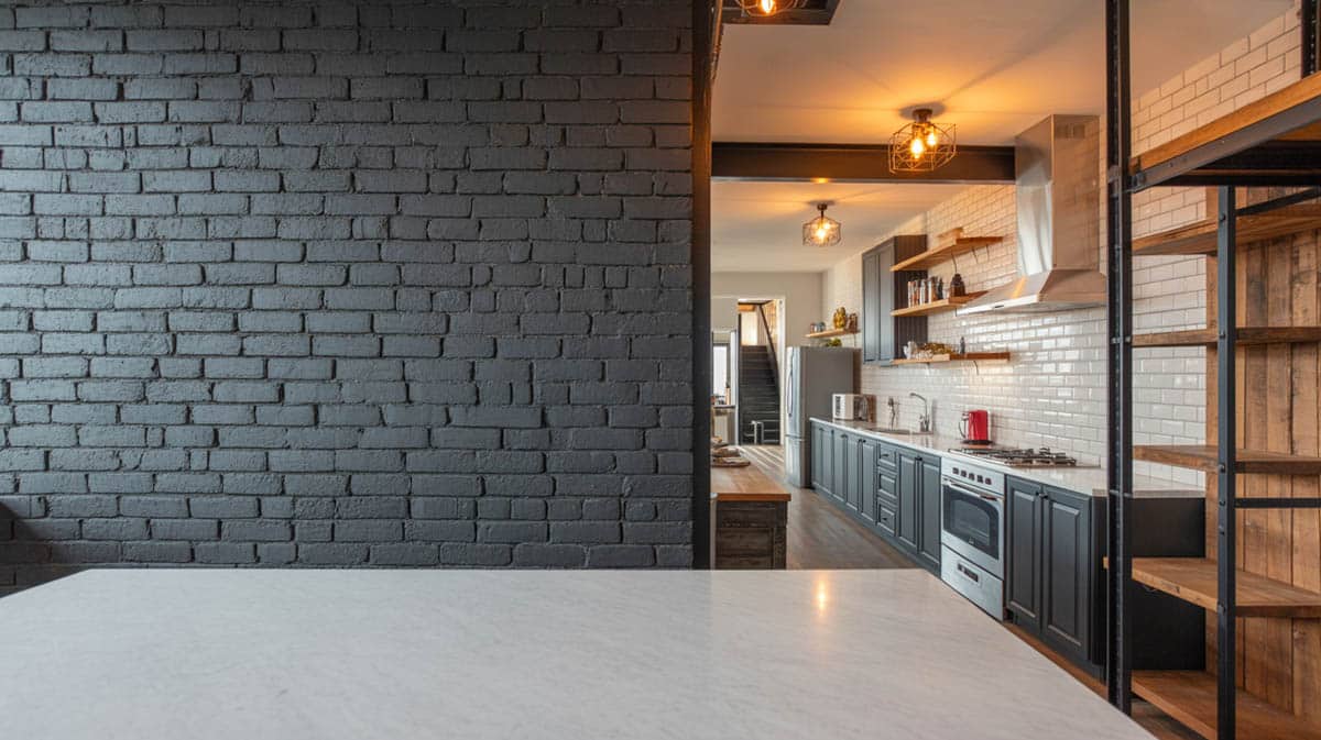 Exposed brick wall painted matte black behind white marble counter with open kitchen view beyond