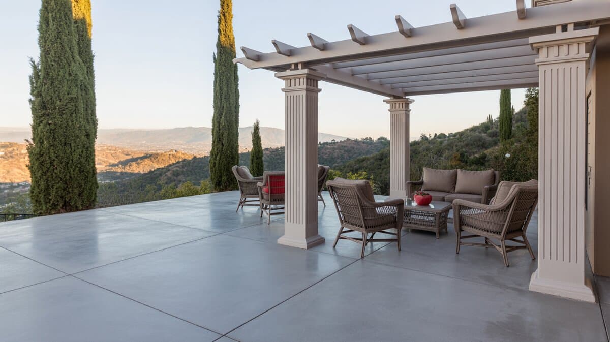 Smooth concrete patio under a classical pergola with fluted columns, wicker seating, Italian cypress trees, and mountain valley views
