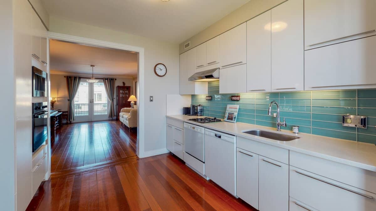 Small white galley kitchen open to living area with aqua glass backsplash and cherry hardwood floors