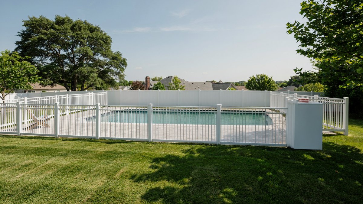 White vinyl safety fence with vertical pickets bordering a green backyard pool
