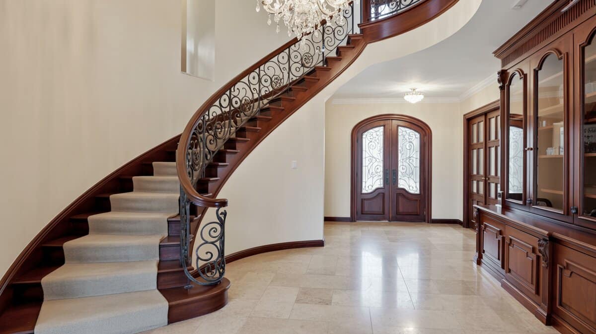 Grand curved staircase with ornate wrought iron scrollwork balusters and crystal chandelier in travertine marble foyer