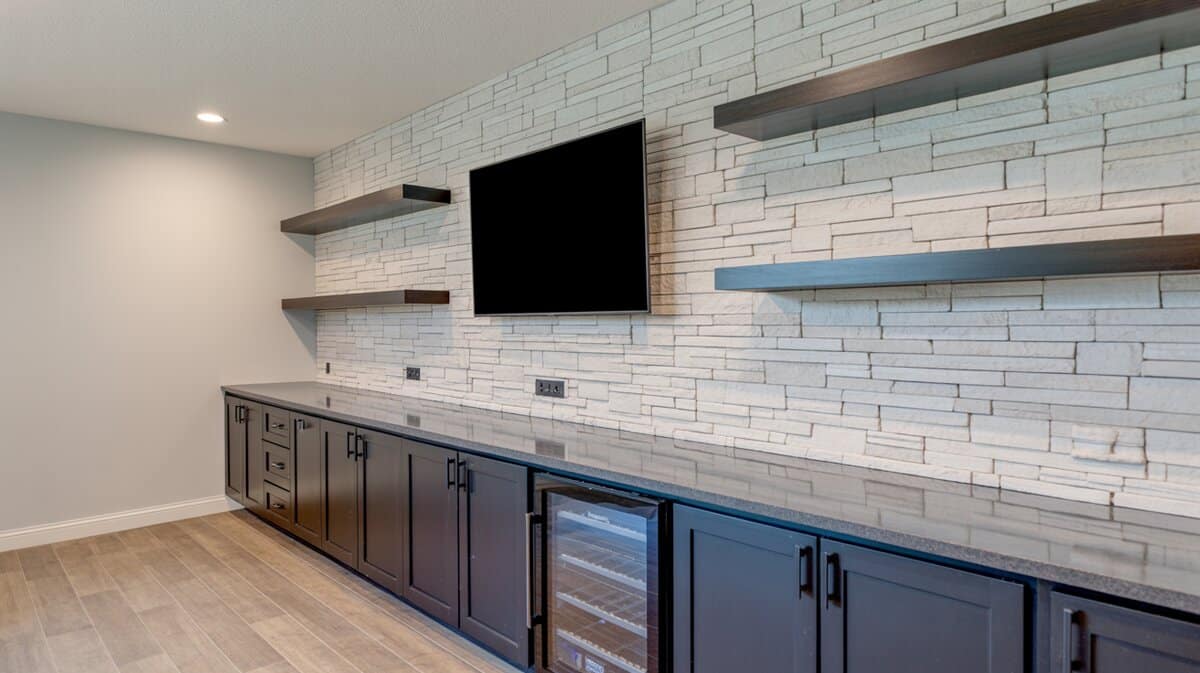 Basement bar with pale stacked stone back wall, dark navy cabinets, and floating shelves