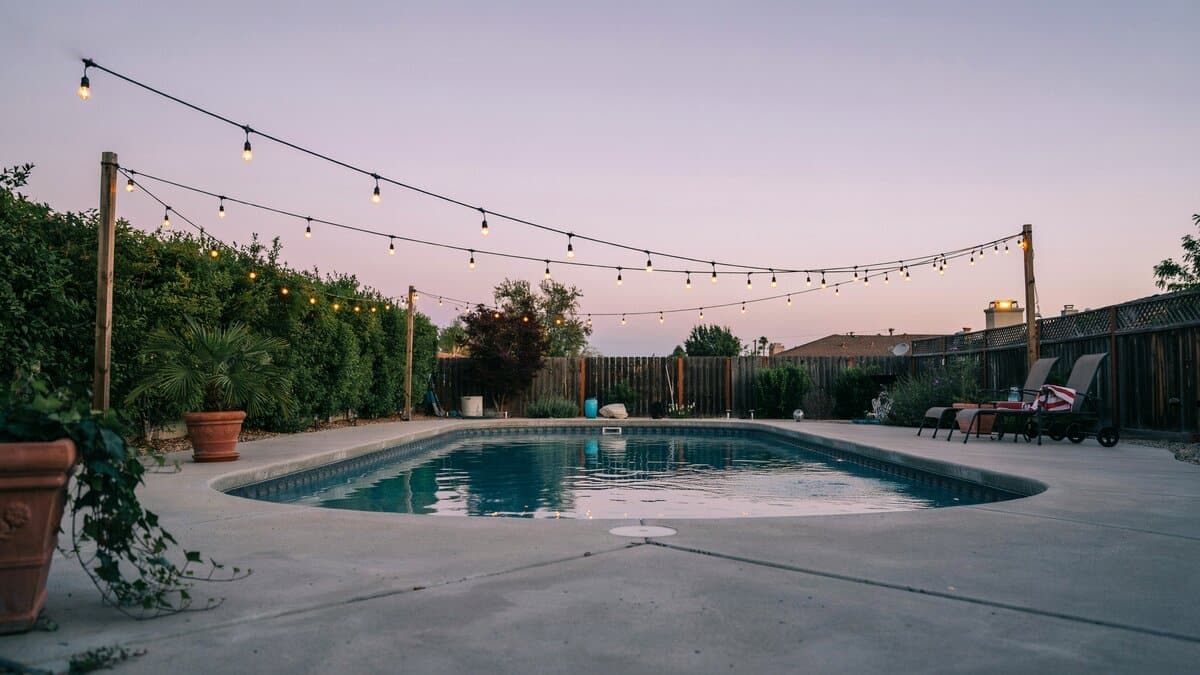 Backyard swimming pool edged with string lights mounted on poles at sunset