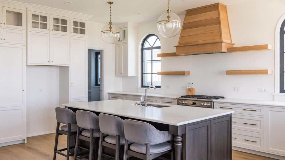 Two-tone kitchen with white quartz island, natural wood range hood, arched window, and gray upholstered bar stools