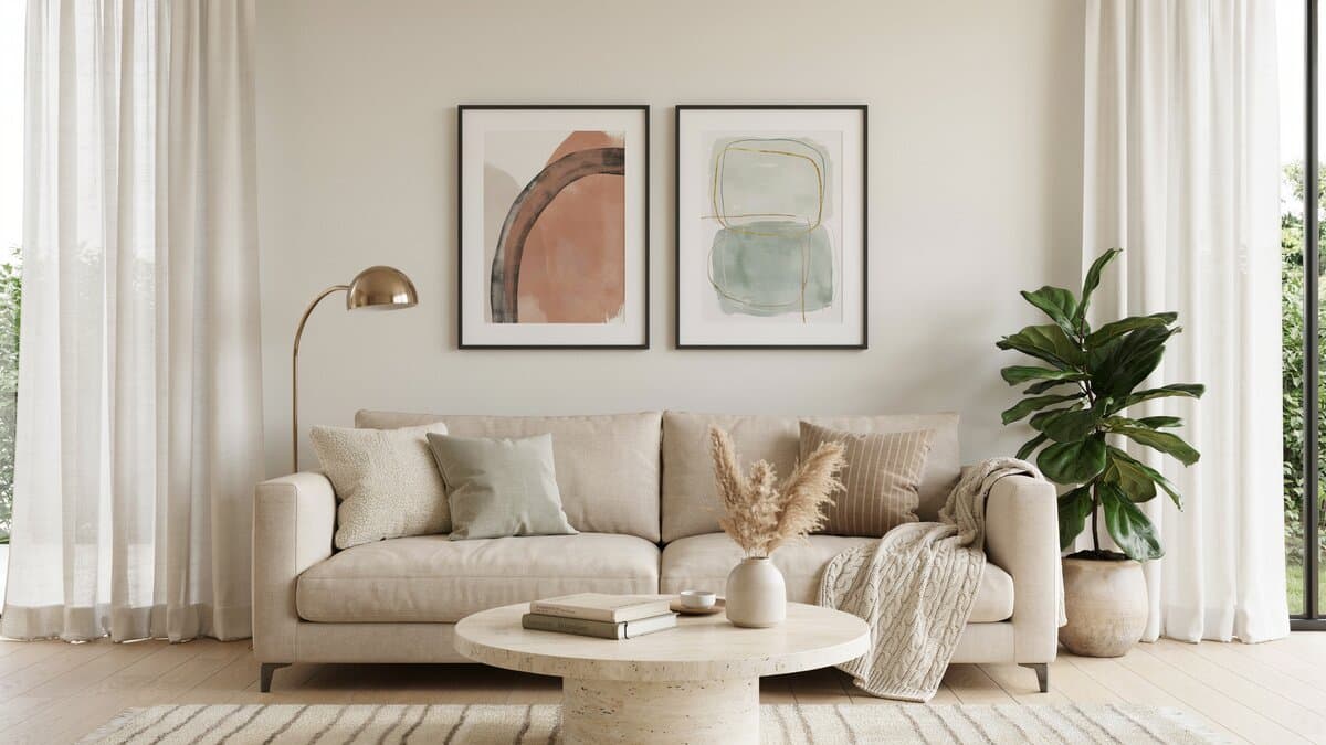 Two large abstract art prints hung above a beige sofa near windows