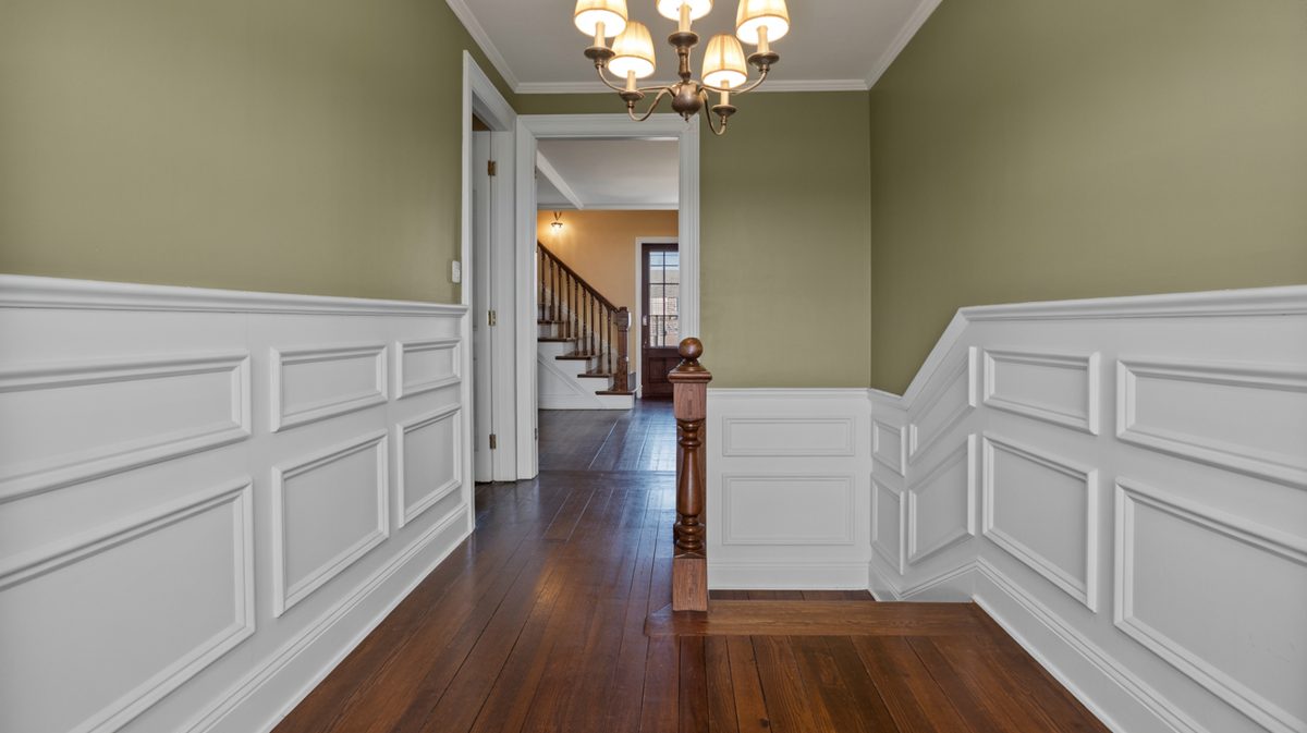 Upper hallway landing with white raised panel wainscoting, sage green walls, oak stair banister, dark hardwood floors, and candelabra chandelier