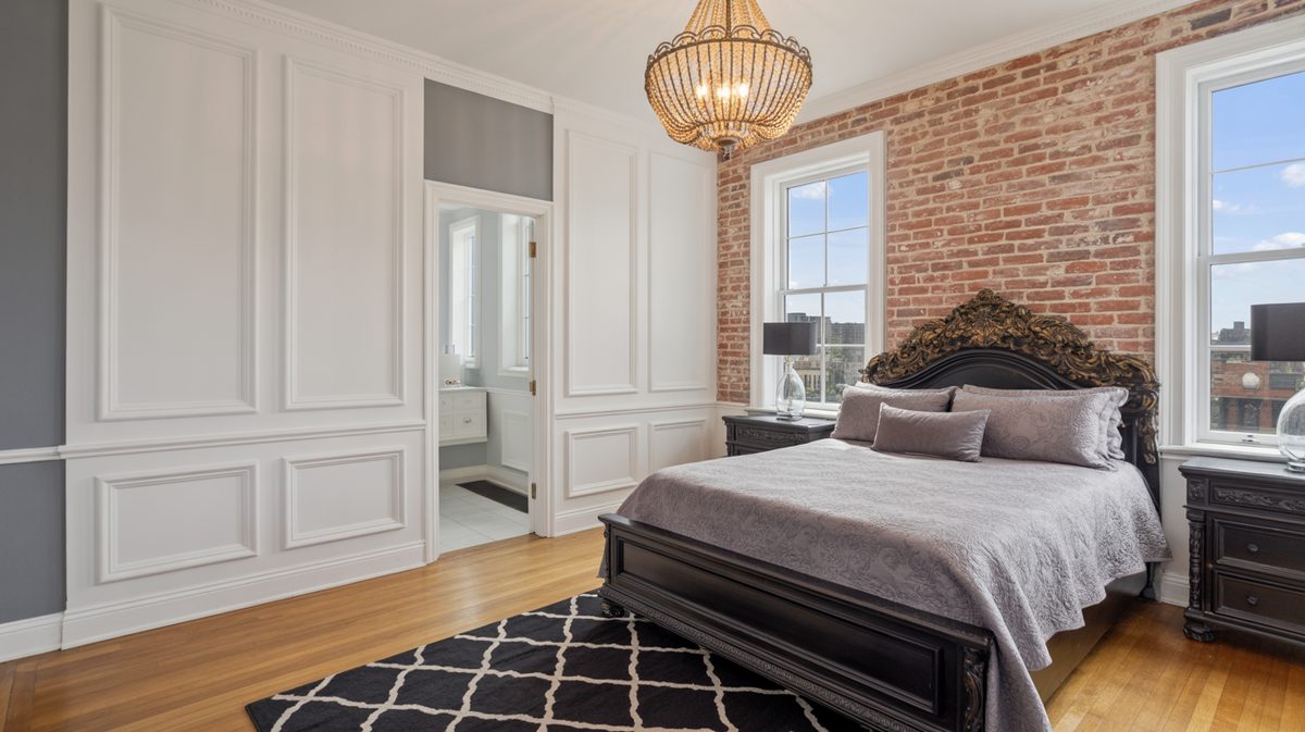 Bedroom with white raised panel wall treatment, exposed red brick accent wall, ornate black carved bed, and gold beaded chandelier overhead