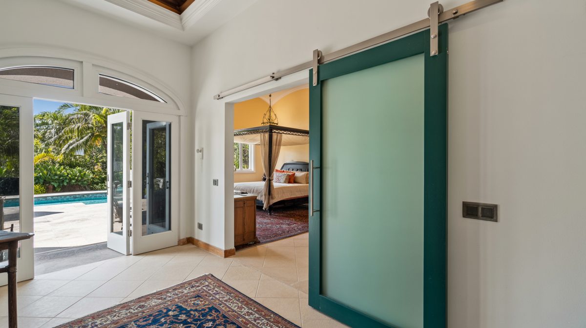 Teal-framed frosted glass sliding barn door on steel rail in a Mediterranean-style home with arched French doors opening to a pool and tropical garden
