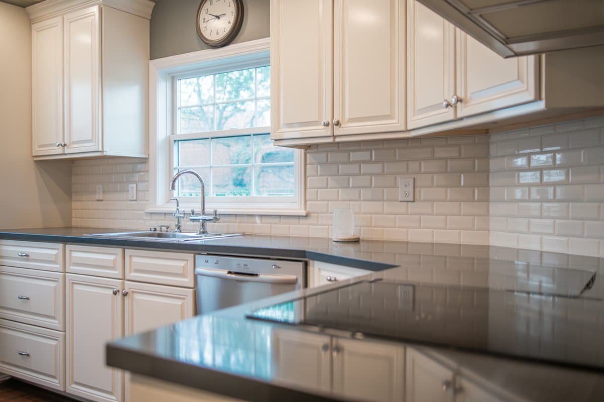Wall clock above a sink window in a white kitchen with dark counters