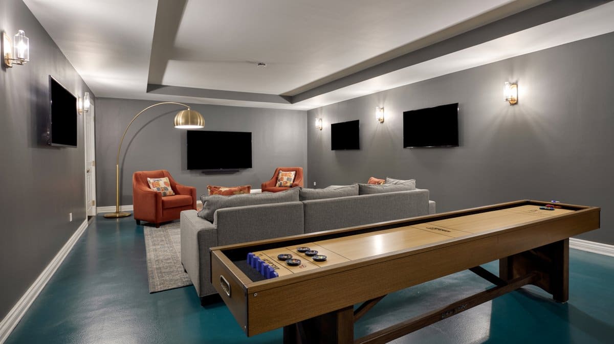 Contemporary gray basement with long shuffleboard table, gray sectional sofa, orange accent chairs, and three wall-mounted TVs
