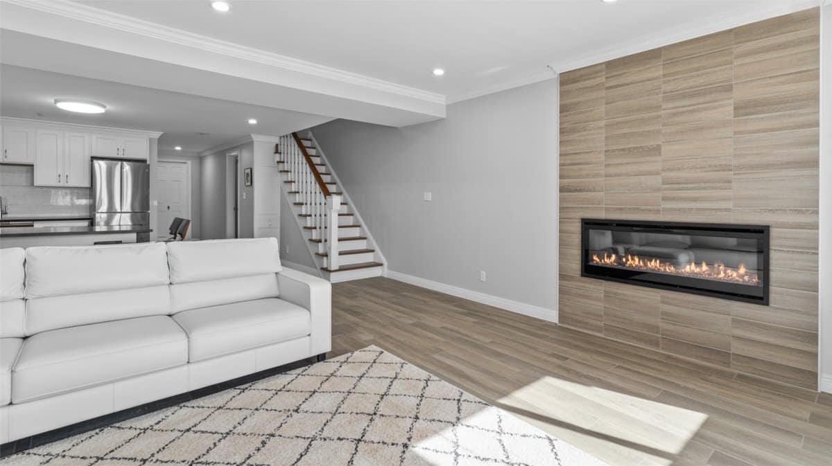 Clean open-plan finished basement with white sofa, horizontal tile fireplace surround, and white kitchen beyond staircase