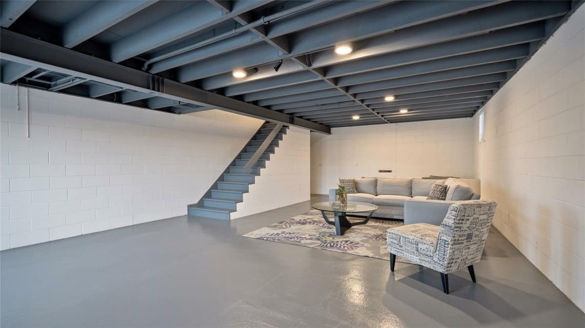 White painted concrete block basement with gray open ceiling, gray sofa, patterned accent chair, and glass coffee table