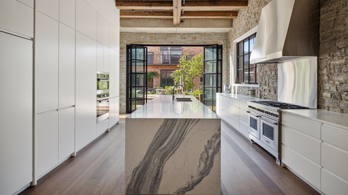 Modern loft kitchen with white cabinets and long marble waterfall island opening to courtyard