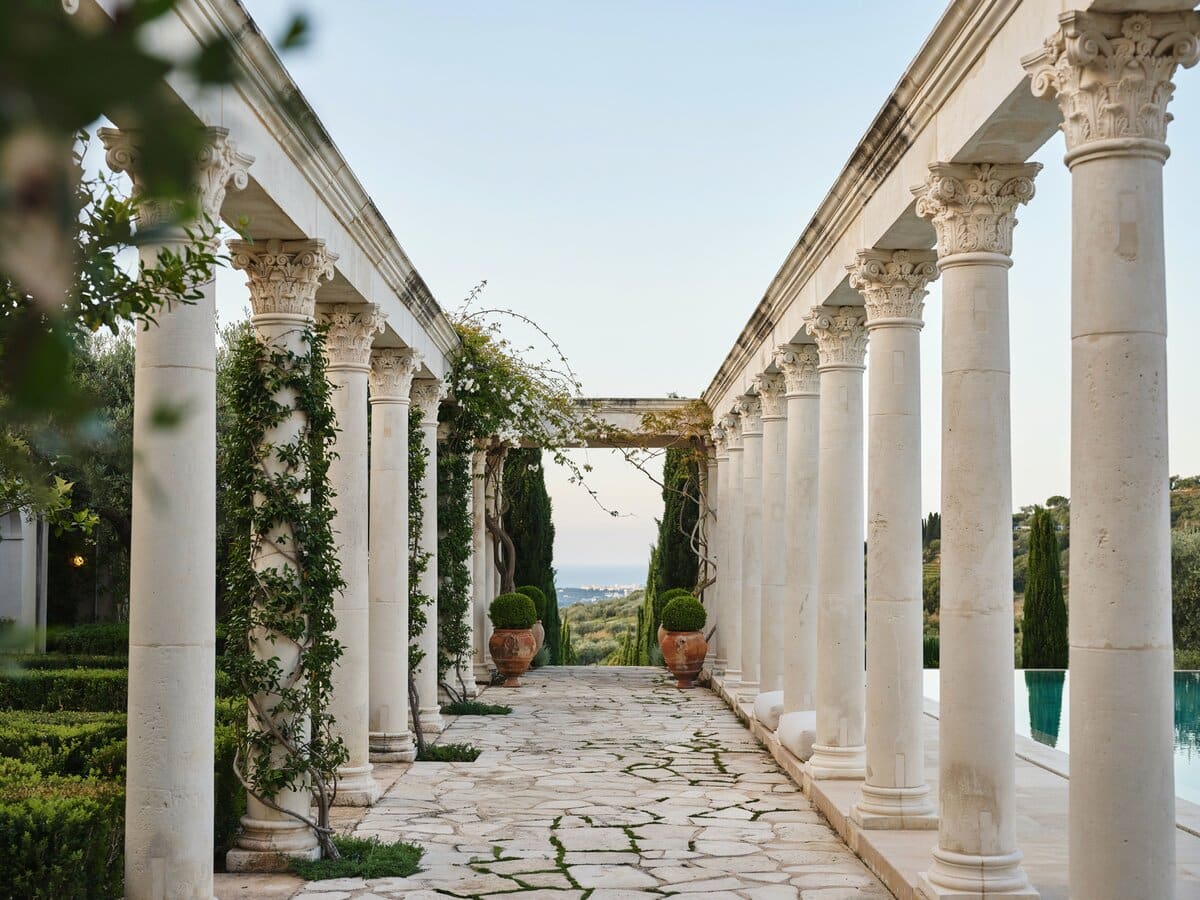 Narrow flagstone walkway between white stone colonnades draped with vines beside a swimming pool