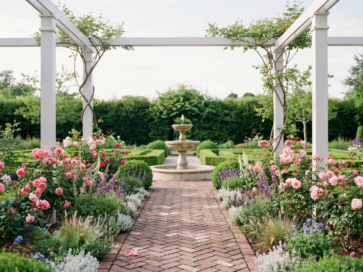 Herringbone brick path under a white pergola leading to a garden fountain with pink flowers