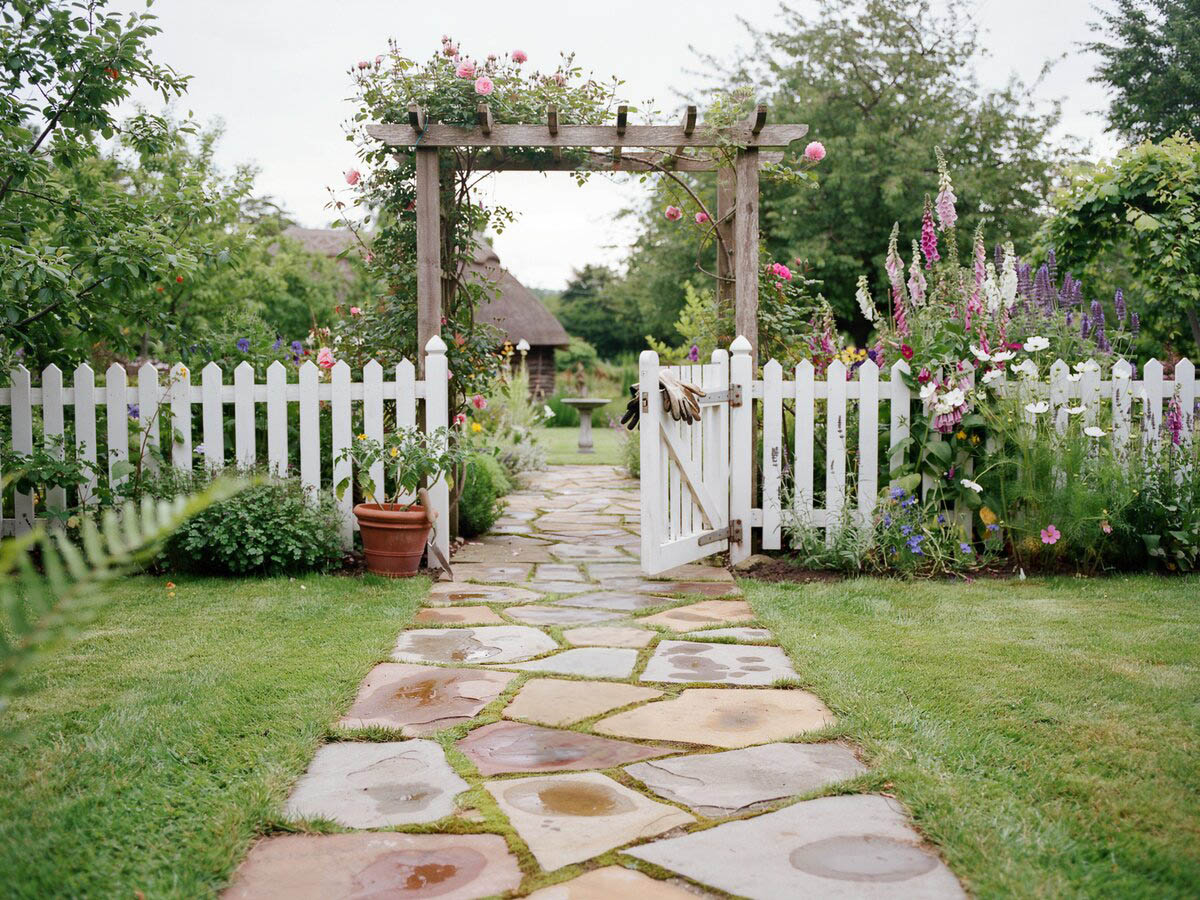 Irregular sandstone flagstone path leading to a wooden arbor garden gate with a white picket fence