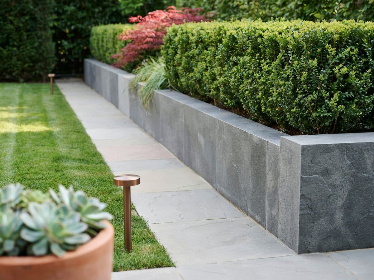 Modern narrow stone tile walkway beside a granite retaining wall with a formal hedgerow