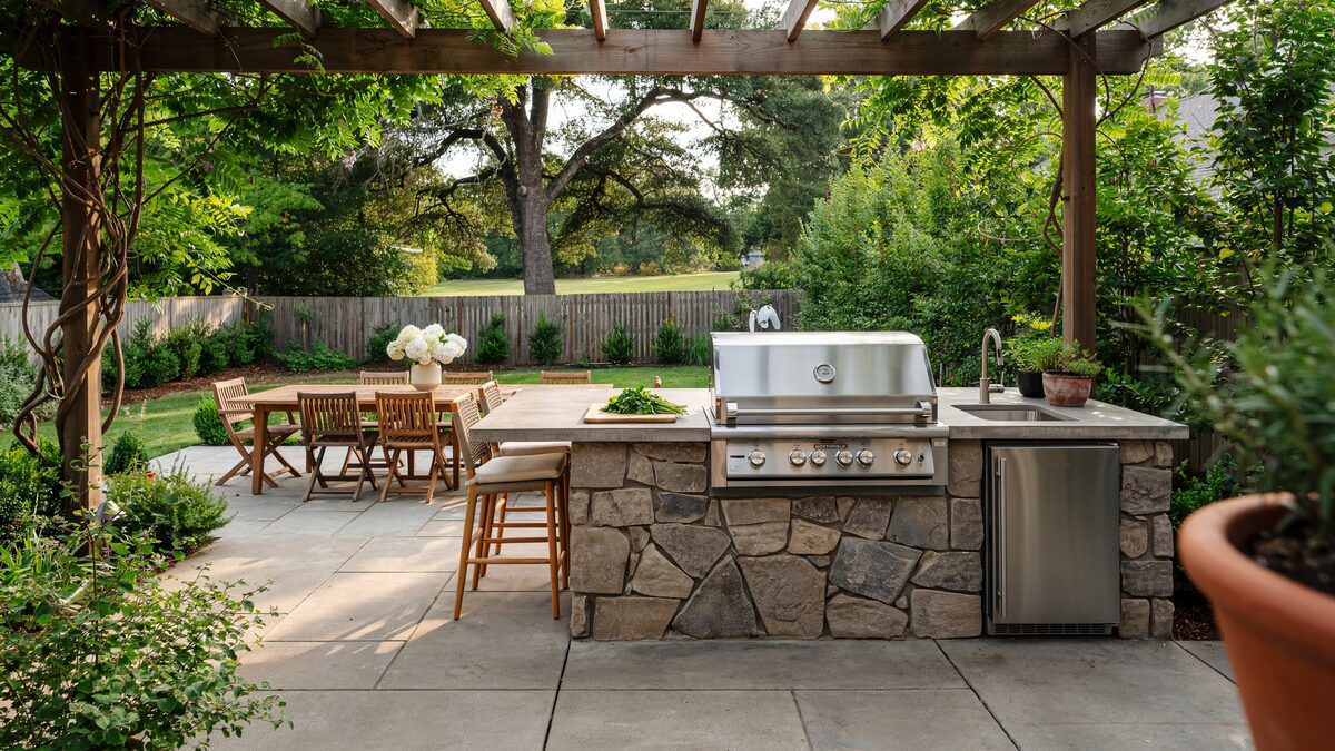Stone-base outdoor kitchen with stainless grill beside a pergola dining area
