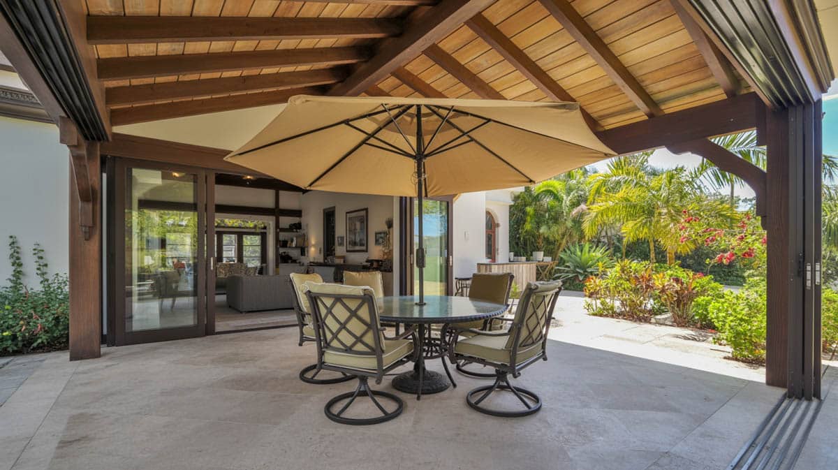 Covered outdoor patio with wood beam ceiling, tan market umbrella, round dining table, and tropical garden