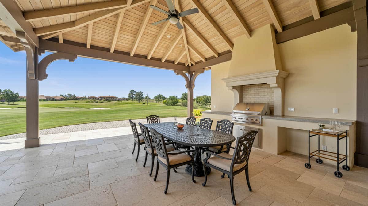 Covered outdoor loggia with vaulted wood beam ceiling, cast iron dining set, outdoor kitchen, and travertine tile