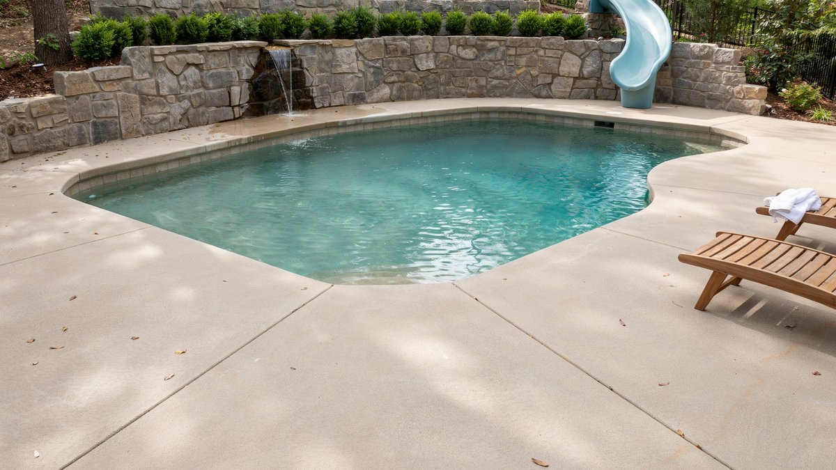 Freeform pool with stacked stone waterfall wall, blue curved slide and broad concrete patio