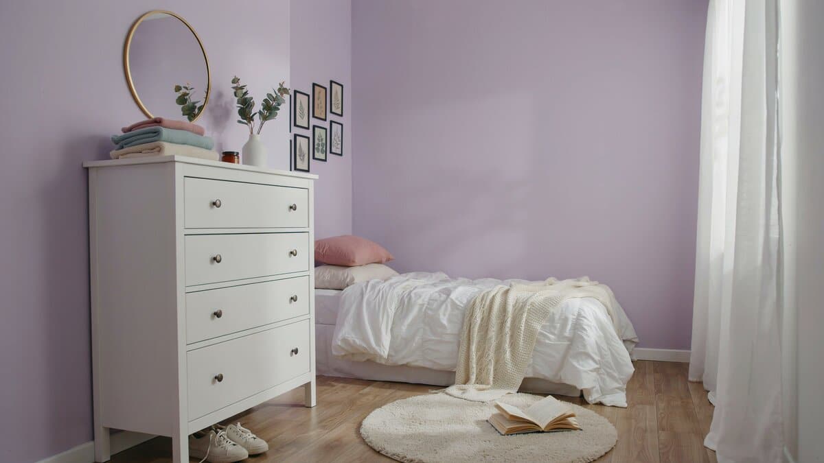 Lavender walls with a white six drawer dresser and round gold mirror