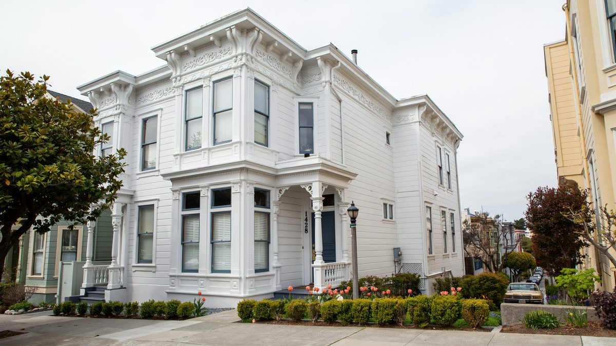 White Victorian corner house with stacked bay windows and landscaped garden beds